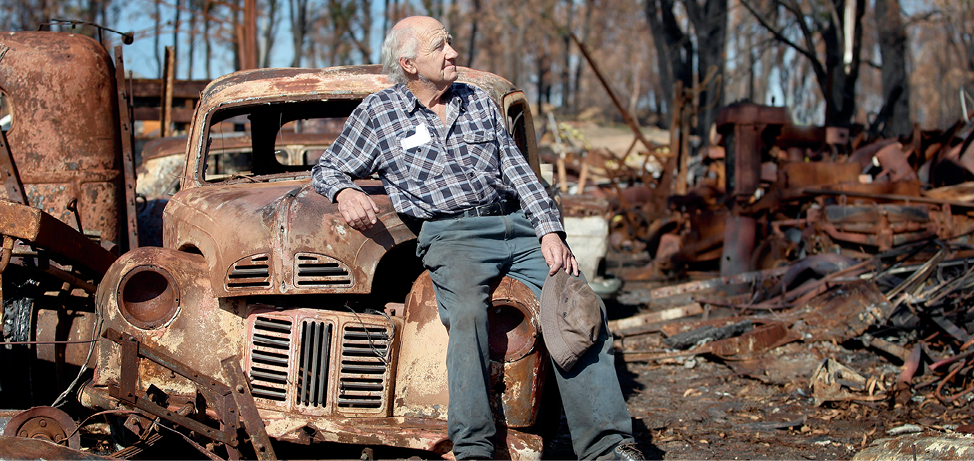 Photograph of Gerald Egan leaning on a burnt-out vehicle with burnt trees and remains of property in the background