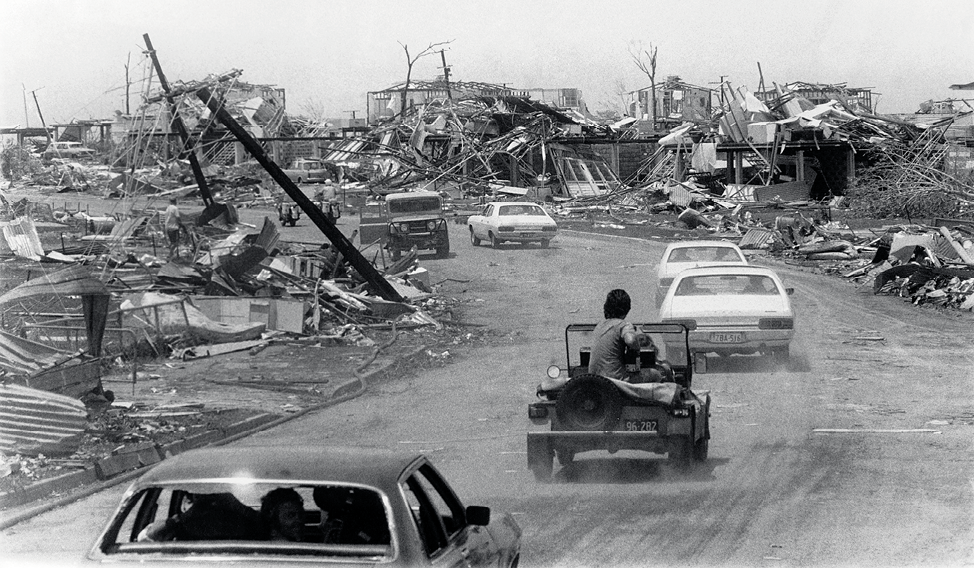 Photograph of a convoy of cars driving through an area with destroyed buildings