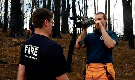 Photograph of one young person filming another outdoors, both wearing Fire services uniforms.