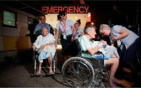 Photograph of two older people in wheelchairs outside a hospital emergency department being assisted by four hospital staff