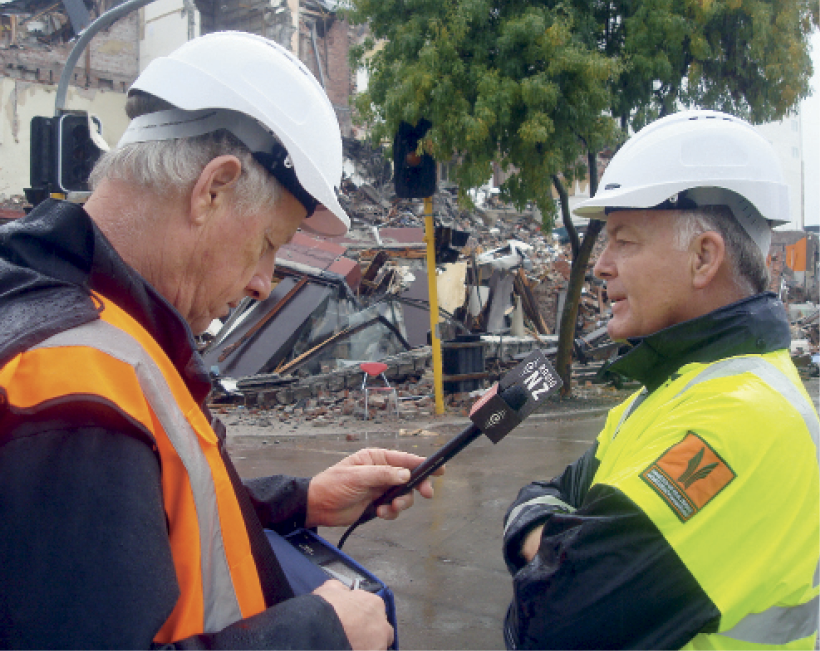 Photograph of John Hamilton being interviewed in a street with destroyed structures in the background