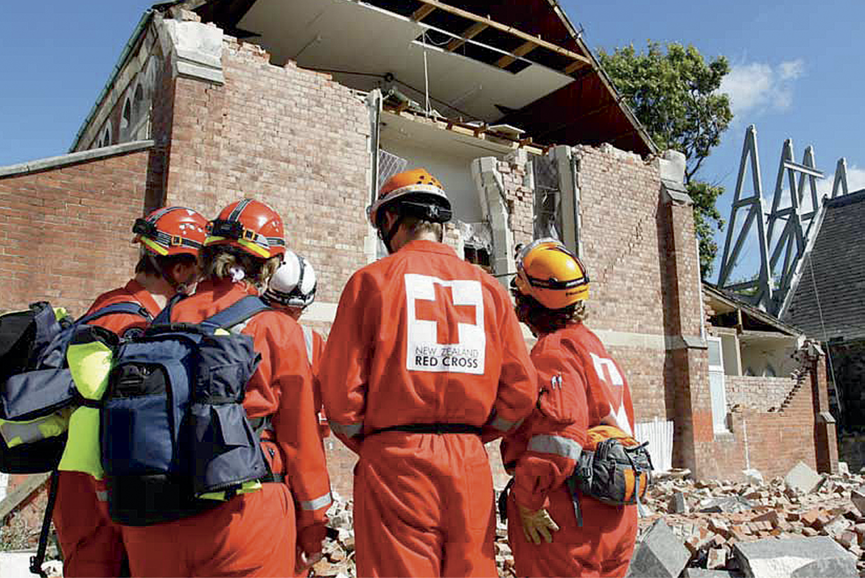 Photograph of team members in Red Cross uniforms and hard hats outside a damaged building
