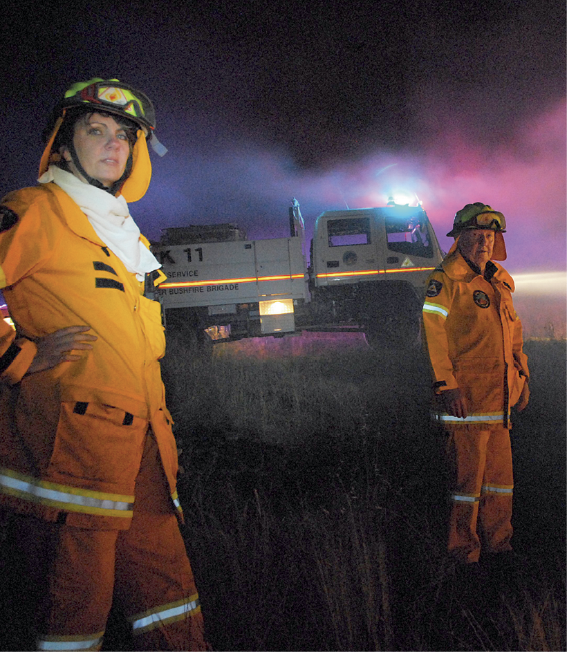 Photograph of a young woman and an older man in emergency services uniforms, with a fire truck and smoke in the background