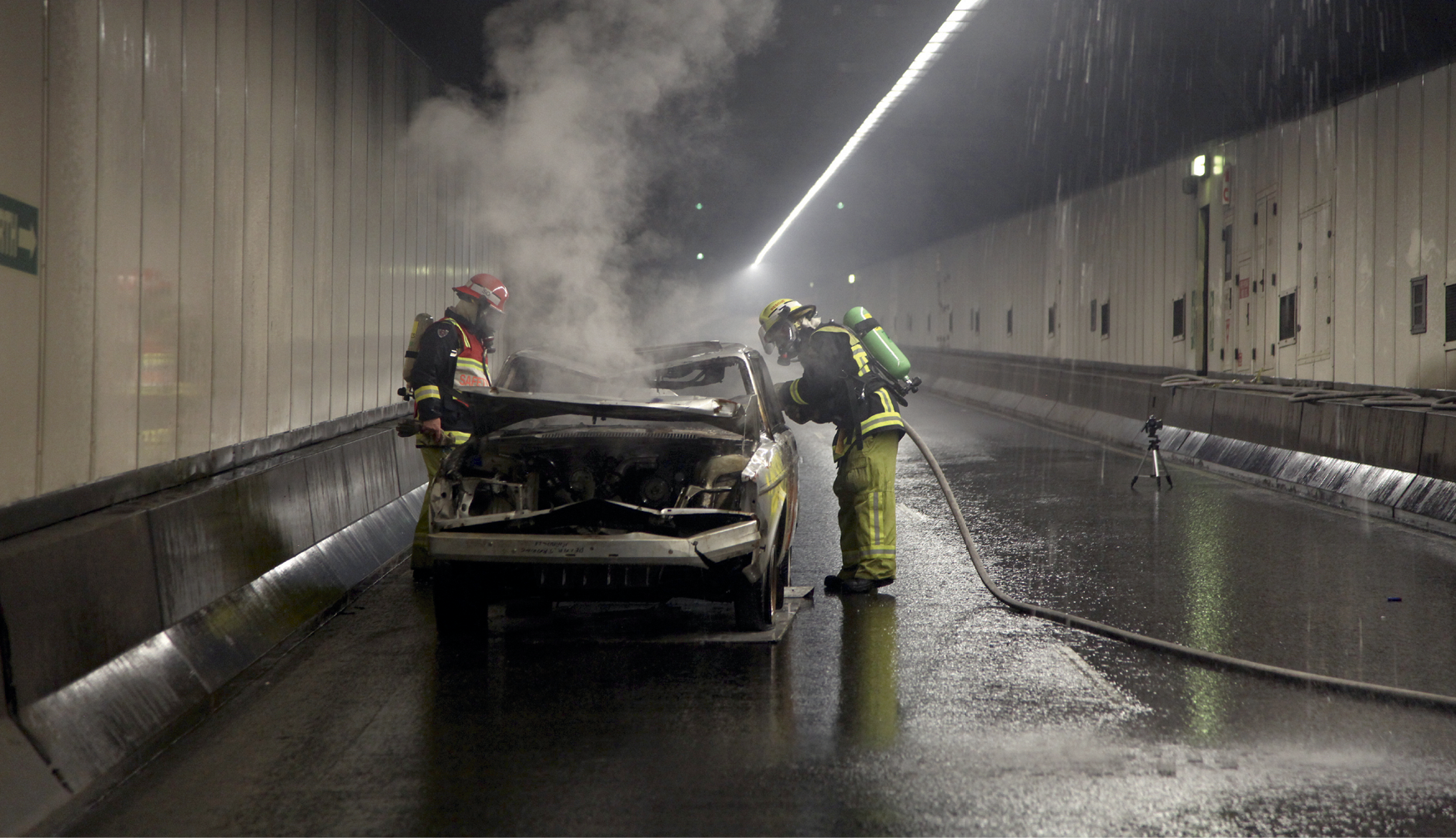 Two firefighters are hosing a burnt-out car inside a road tunnel. There is smoke rising from the bonnet of the car and water spraying from the roof of the tunnel.