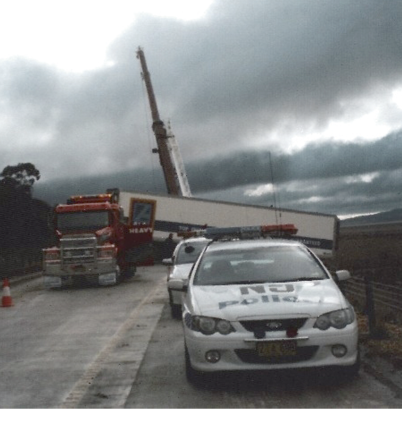 In the foreground are two police cars. In the background is a semi-trailer truck with its trailer at a right angle across the road and through the roadside barrier. Behind the truck is the top of a crane arm.