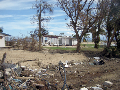 In the background are the standing remains of a partially destroyed single-storey house. In the foreground are piles of debris.