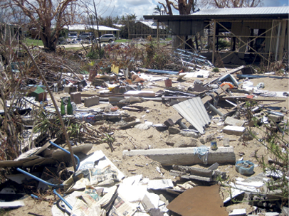 A yard is covered in debris including a variety of building materials. In the background is the standing remains of a single-storey house.