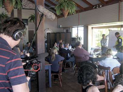 Many adults are seated in a vaulted dining room. They are facing a man in the background who is writing on a large flipchart. In the foreground is a man operating a television camera.