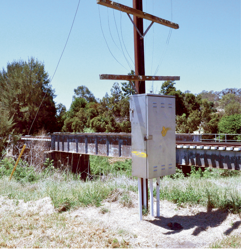 An electrical box on a post has a yellow paint line one third of the way up the box, approximately level with the railway bridge beside it.