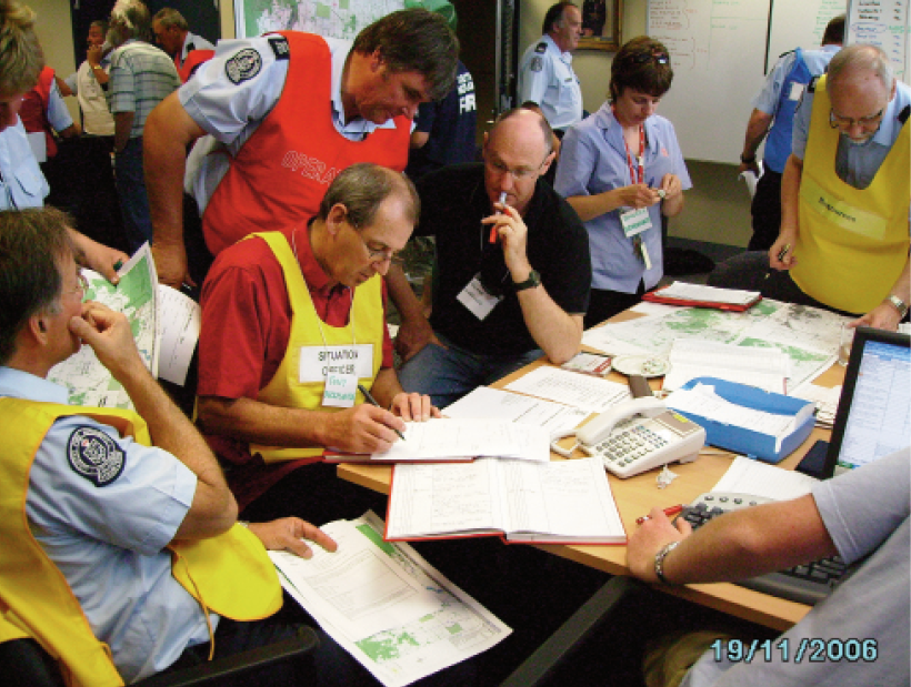Several male and female emergency services personnel are gathered around a table covered in papers and maps