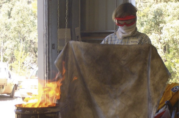 A woman wearing a protective face wrap and goggles is about to put out a small fire in a metal drum using a large cloth