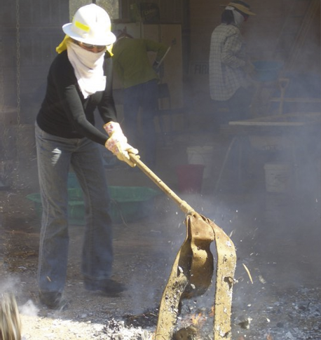 A woman wearing jeans, long-sleeve shirt, protective gloves, face-wrap, hard hat and glasses is putting out a small fire using a leather strap on a long handle.
