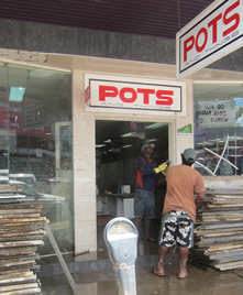 A man wearing rubber gloves is cleaning the doorway of a Pots shop front. Both the floor inside the shop and the ground outside the shop are wet.
