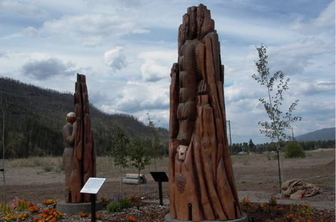 Two carved wooden monuments stand in a small garden bed surrounded by barren plain. In the background are hills covered with burnt pine trees.