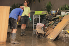 A man and a woman dressed in mud-splattered shorts, t-shirts, gumboots and rubber gloves are stacking ruined furniture and household items in a driveway and clearing mud from the driveway.