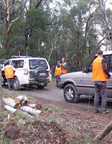 Two 4-wheel drive vehicles are parked on a bush road beside a wire fence with bushland in the background. Several people dressed in orange safety bibs are standing around the vehicles. One man is using a thermal imaging cameral to look up into the trees beside the road.