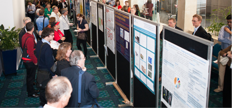 A crowd of people are looking at stands displaying information posters in a long wide hallway.