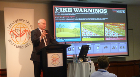 Victorian Fire Services Commissioner, Craig Lapsley is speaking from a lectern in front of a seated audience with a large screen in the background displaying images and text about fire warnings.