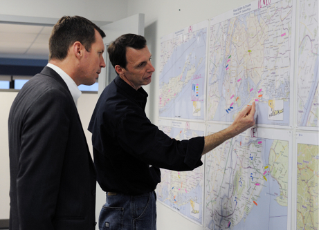 Timothy Manning and another man examine several maps on the wall, on which different sites are marked.
