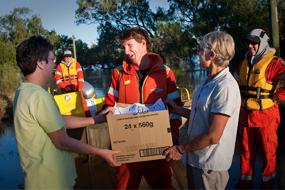 Five people are shown by the bank of a flooded river. Two of the people are receiving a food parcel from an emergency services volunteer, while another two emergency services personnel stand on in the background.