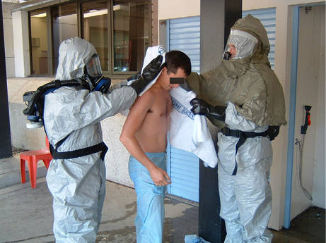 Two people wearing breathing apparatus and biohazard suits practice decontaminating a practice subject.