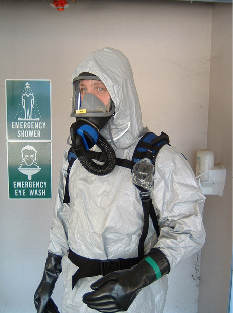 A person wearing breathing apparatus and a biohazard suit, standing under an emergency shower.
