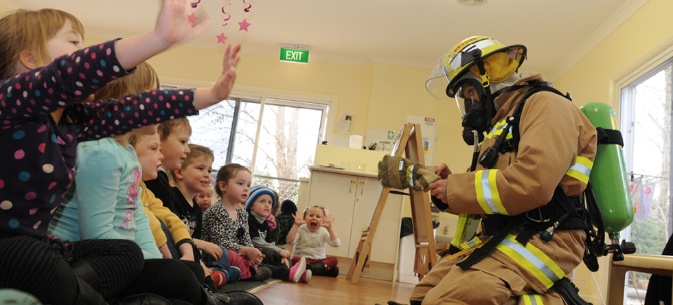 A photo of a firefighter kneeling in front of several children indoors, showing them fire-safe clothing and equipment.