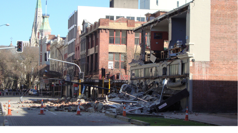 A photo of a street in Christchurch after an earthquake. It shows damaged buildings and street fixtures, and rubble that has fallen onto the footpath and road.