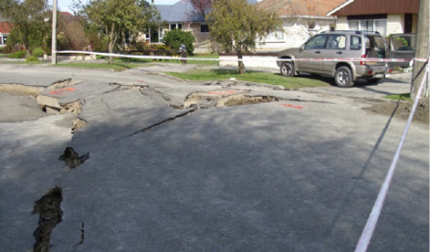 A photo of a damaged street in a residential neighbourhood. The street is broken up and large holes are visible. The area is marked off with tape to prevent people from driving and walking over it.