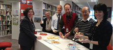 A photo of six people standing at a library counter eating scones. 