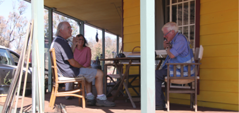 A photo of a male researcher interviewing a man and a woman. They are all sitting outside on the porch of a weatherboard building.