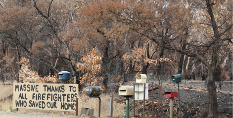 A photo of a hand-painted thank-you sign in front of a group of roadside mailboxes, which reads ‘Massive thanks to all the firefighters who saved our home’. In the background the bush is burnt.