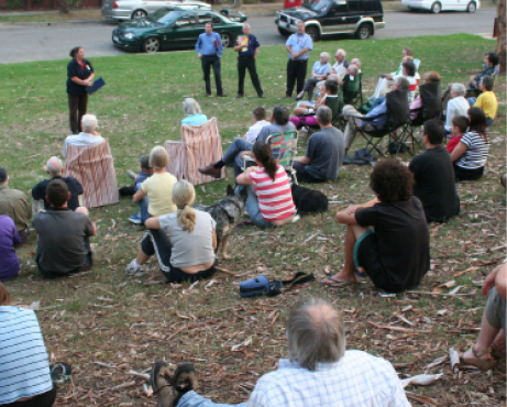 Members of the community are sitting on grass, listening to a presentation.