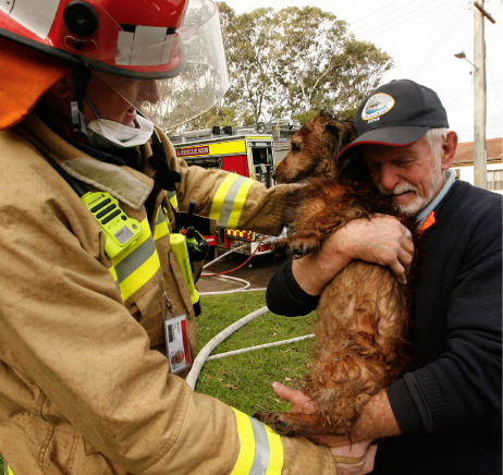 A firefighter is handing a small dog to a middle-aged man who is hugging the dog.