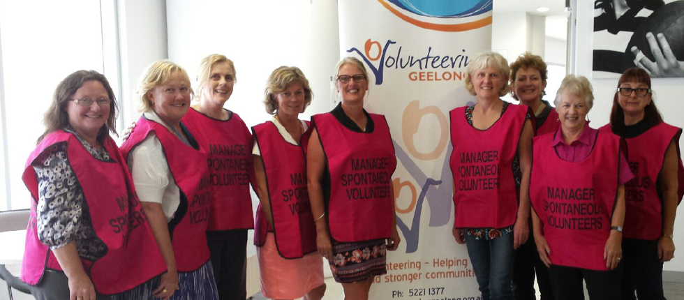 Nine women, all wearing pink ‘Manager spontaneous volunteers’ vests, are standing in front of a Volunteering Geelong banner.
