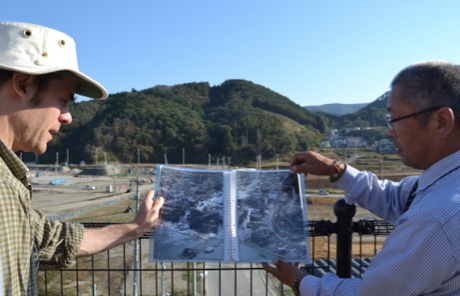 A caucasian man and a Japanese man are comparing a photo of a flooded landscape to the same landscape, dry, in the background.