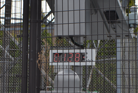 Some grey electronic equipment with a large dosimeter digital display is enclosed by security fencing. 