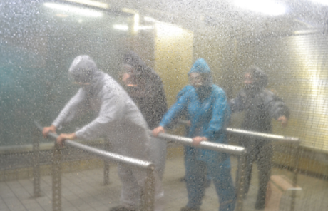 Four people dressed in coveralls, hoods and face masks are holding handrails in a tiled room with a glass observation wall. The room is filled with spraying water and the people are braced against strong wind.