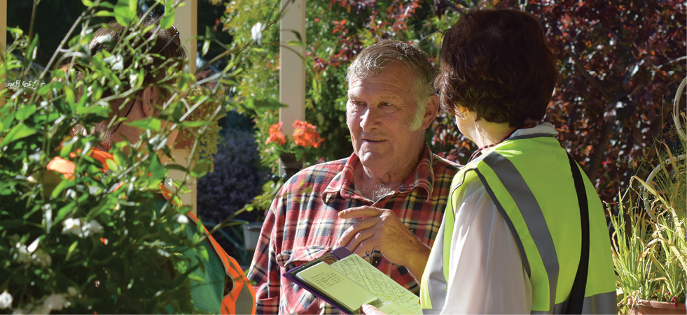 Two women wearing safety vests and holding a clipboard are talking with an elderly man in a very leafy garden.
