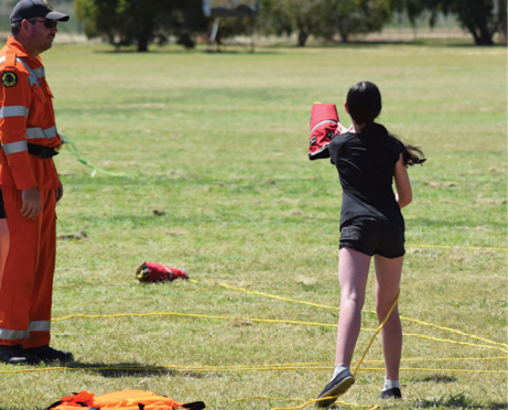 A girl in shorts and t-shirt is practicing throwing a weighted bag attached to a long rope across a grass field. She is being observed by an emergency services man.