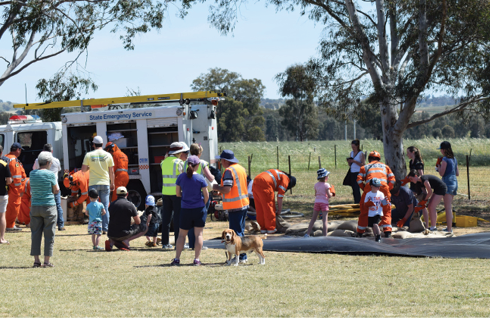 A number of adults, children, dogs and emergency services personnel are engaged in a variety of training activities around an emergency services equipment vehicle.