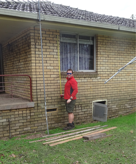 A man is standing in front of a brick house showing that the flood water had reached almost to the eave. The brick walls have some mud still attached.