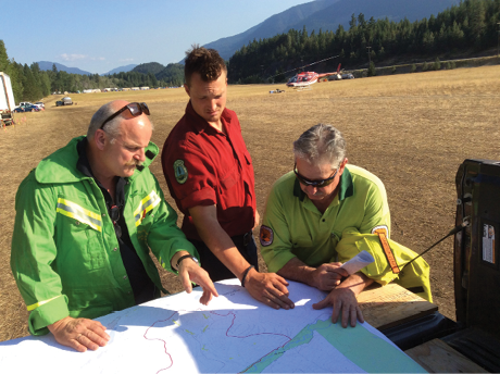 Three men wearing various uniforms are consulting a large map. In the background is a large flat field, a helicopter and pine-forested mountains.