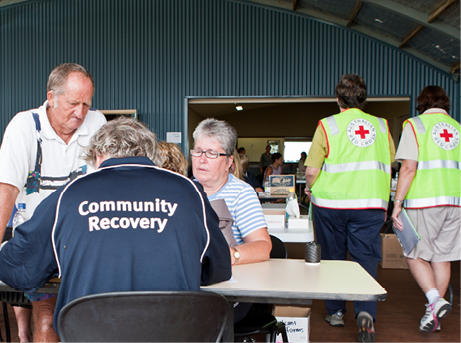 A senior man and woman are consulting with a senior man seated at a table wearing a shirt labelled ‘community recovery’. Beyond them, two adults wearing hi-vis Red Cross vests are walking through a large hall.
