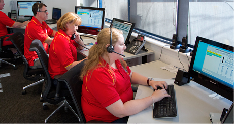 Four adults wearing red polo shirts and headsets are seated at computers in front of a large window.