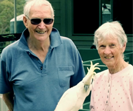 An elderly man and and elderly woman holding a sulphur-crested cockatoo are standing in front of a house with a rainwater tank and bush in the background.