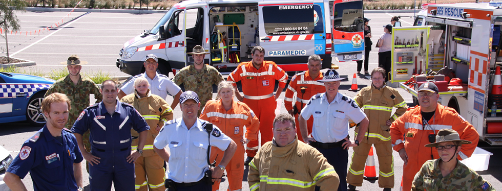 A group of women and men wearing a variety of police, ambulance, defence and emergency services uniforms are standing with emergency services vehicles on display in the background.