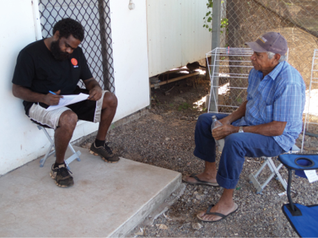 Two indigenous men are sitting outside a simple house. One man is filling in a form.