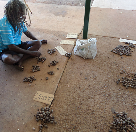 An indigenous man is seated on the ground sorting pebbles into piles with hand-written paper labels including ‘cyclones’ and ‘fires’.