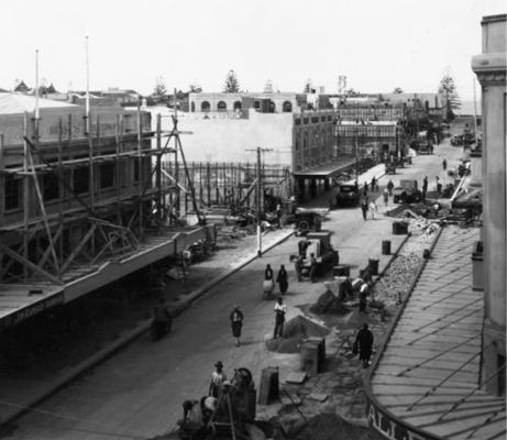A vintage black and white photograph of a main street with most buildings surrounded by scaffolding and piles of materials on the street.